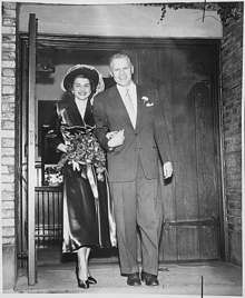 man in a suit leads a flower carrying woman by the hand, walking out 